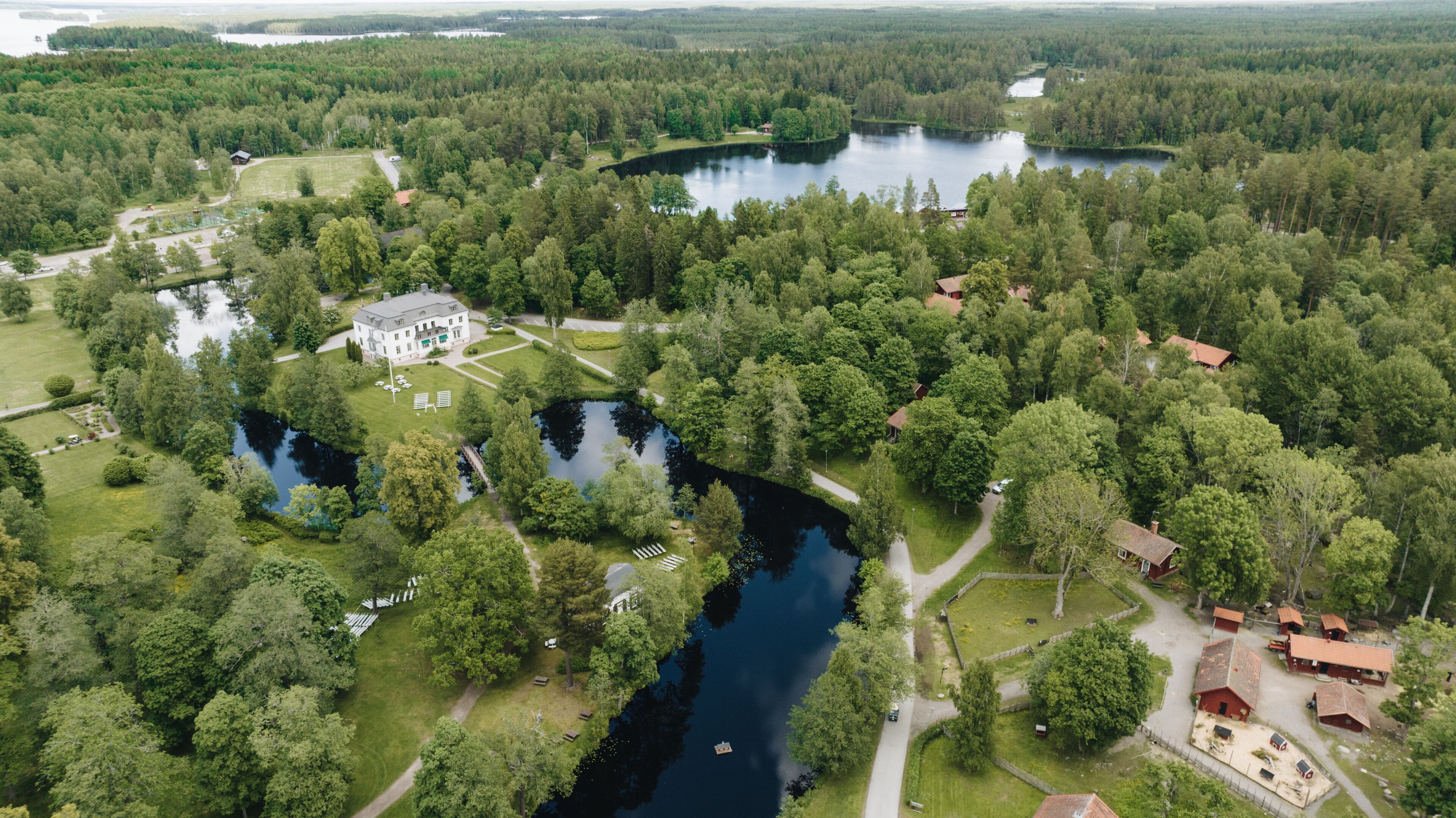 An aerial view of a lakeside property with a large house, multiple smaller buildings, and a well-maintained garden with a pathway leading to the water.