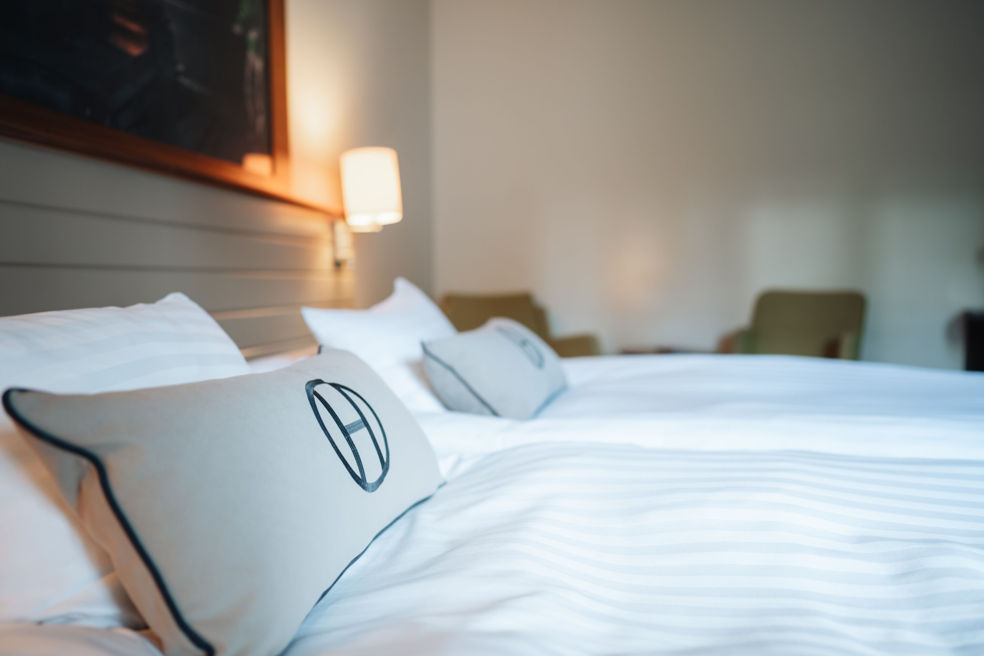 A neatly made bed with white bedding and a pillow featuring a circular logo.