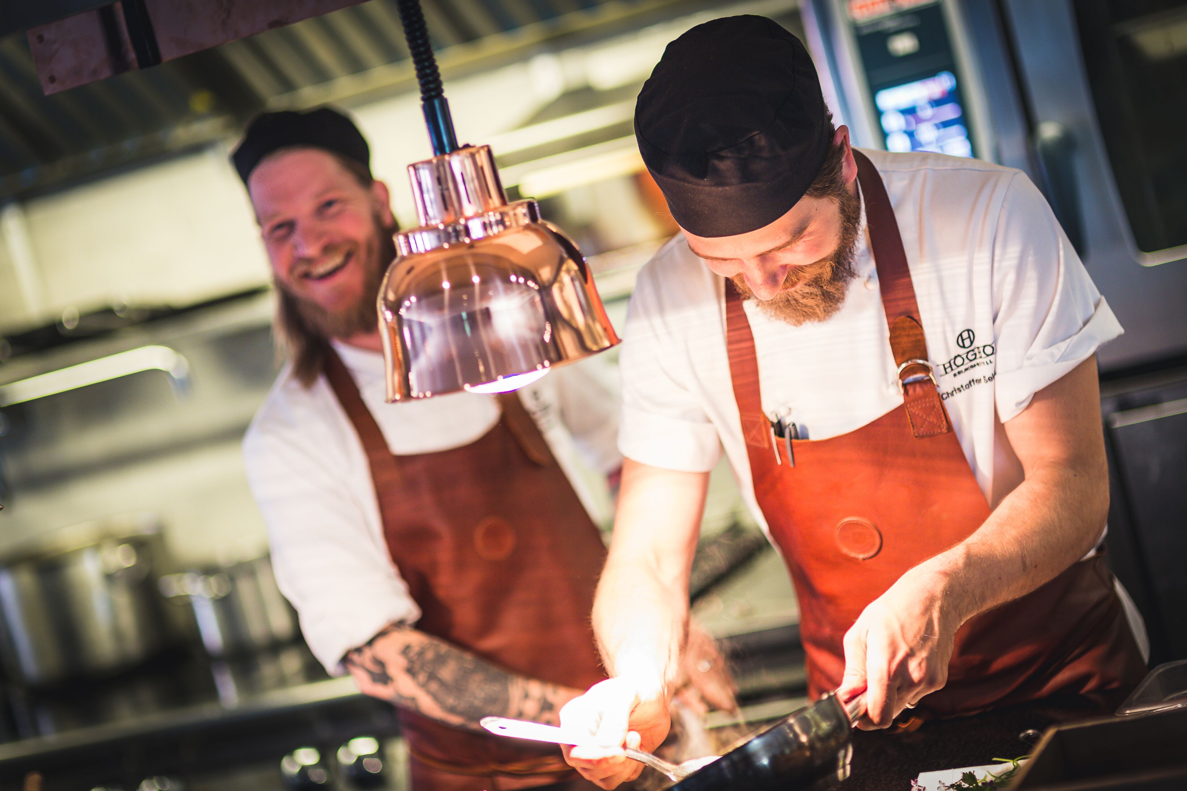 Two chefs in a kitchen, one preparing food and the other smiling.