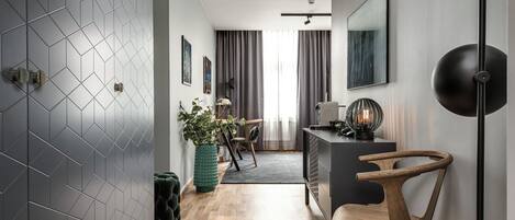 Una sala de estar moderna con un muro de azulejos geométricos, suelo de madera, una planta verde, un sofá negro y un escritorio con una lámpara.