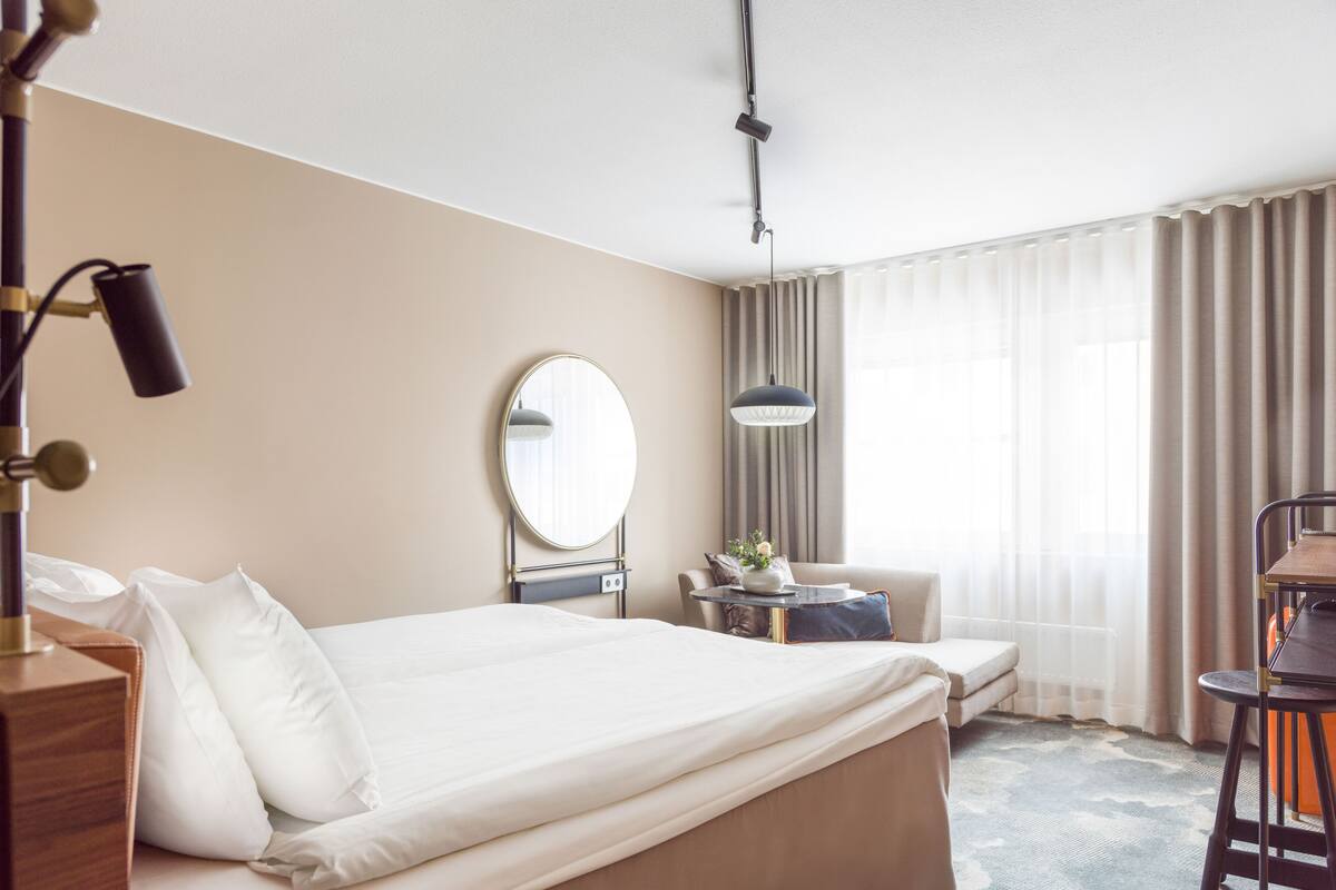 Hotellihuone, jossa on suuri sänky, yöpöytä lampun kanssa, pyöreä peili, pieni pöytä maljakolla, tuoli ja työpöytä tuolilla.