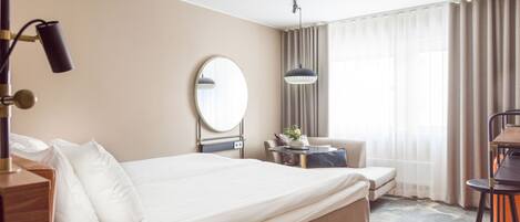 Habitación de hotel con una cama grande, una mesita de noche con lámpara, un espejo redondo, una pequeña mesa con un florero, una silla y un escritorio con silla.