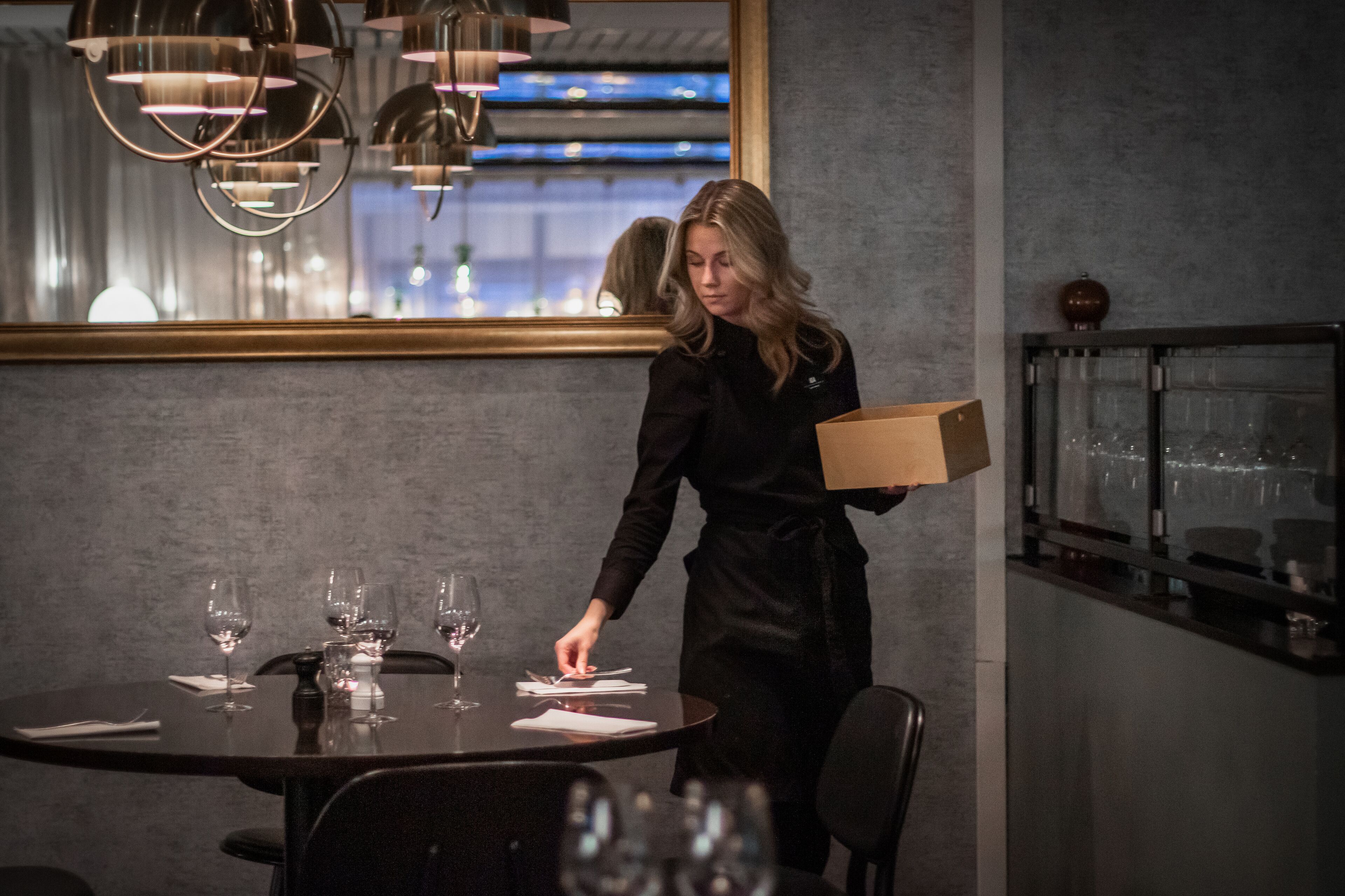 A person in a black uniform carrying a box near a dining table with wine glasses and plates, in a room with a large window and pendant lights.
