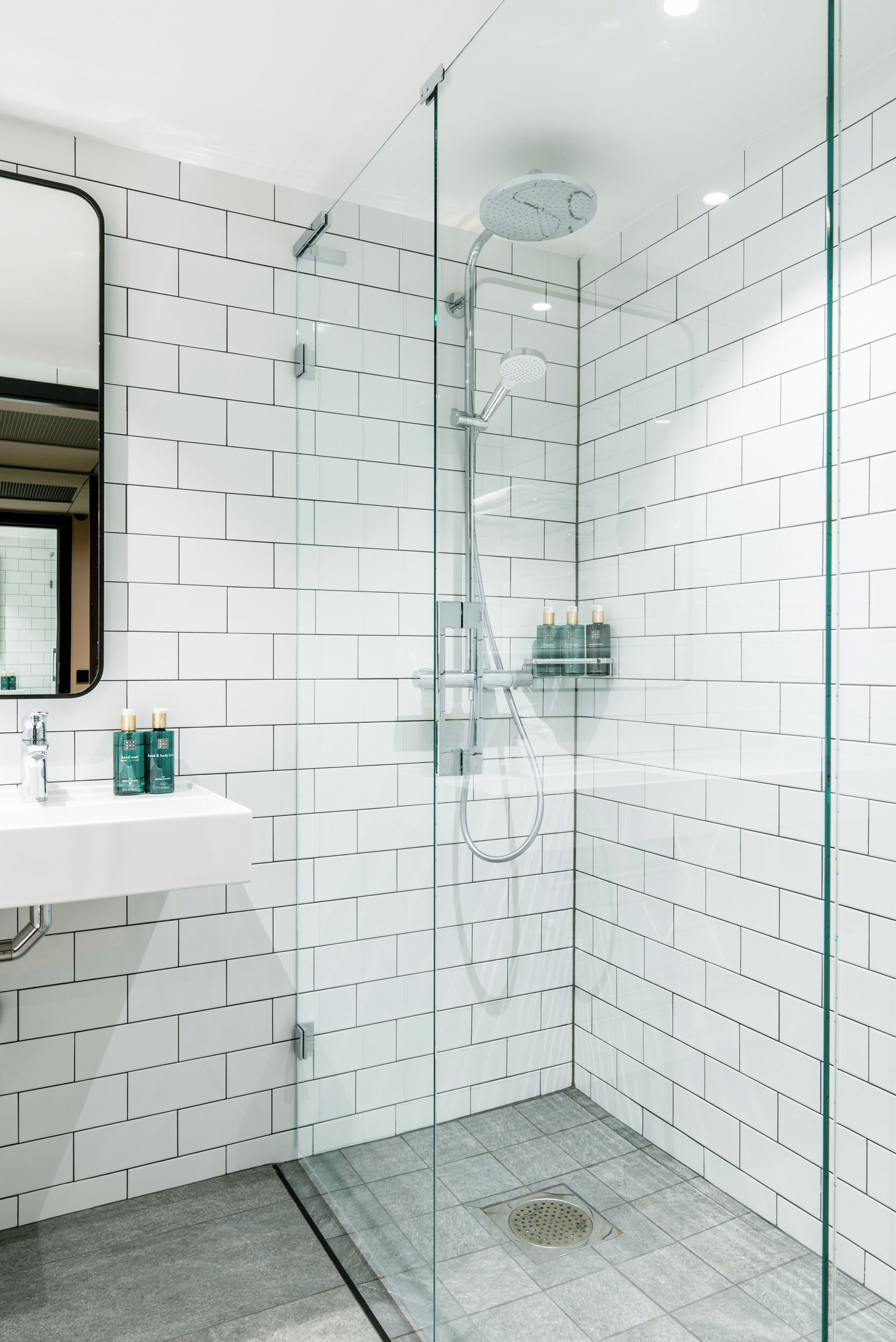 A modern bathroom with white tiles, a glass shower enclosure, and a sink with toiletries.