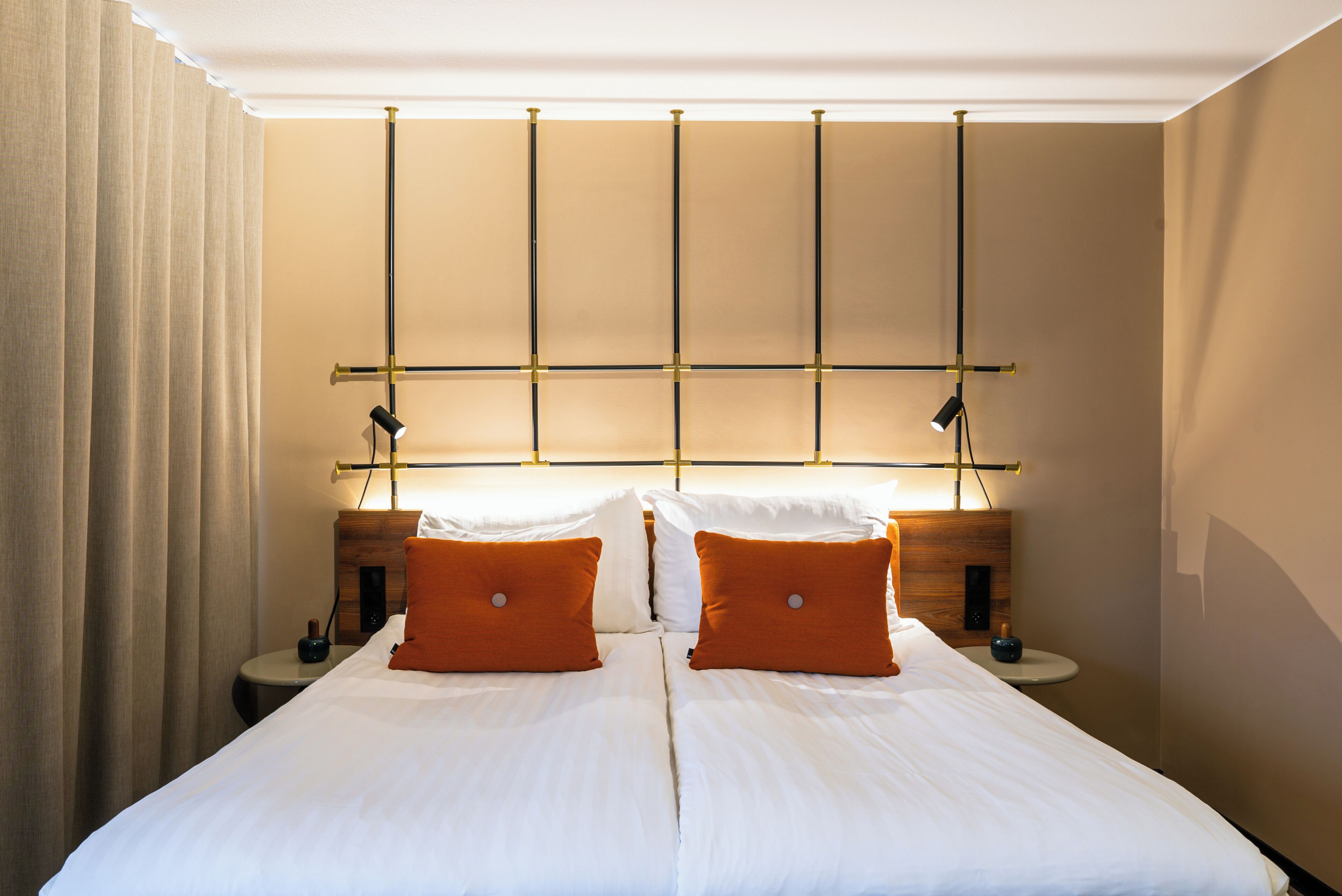 Uma cama de casal com lençóis brancos e duas almofadas laranja, uma mesa de cabeceira com abajur e um painel de cabeceira de madeira com padrão de grade.
