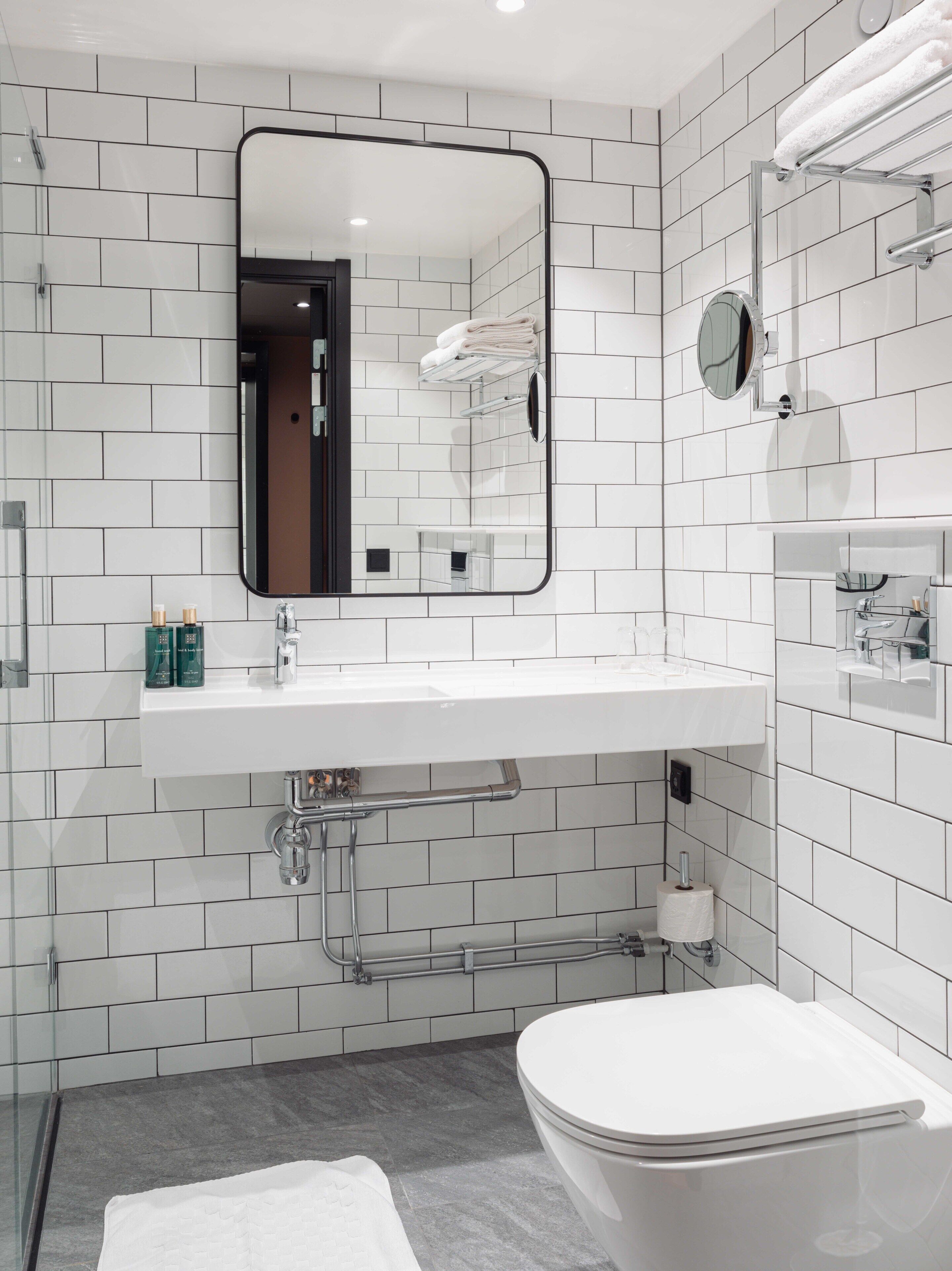 A modern bathroom with white tiled walls, a large mirror, a white sink, and a toilet.