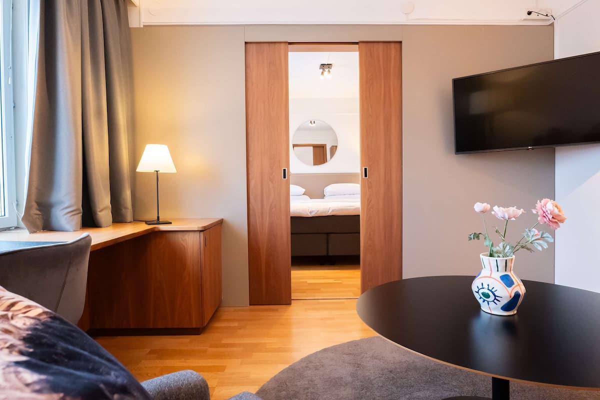 A modern hotel room with a wooden desk, a bedside table, a TV mounted on the wall, and a vase with flowers on a round table.