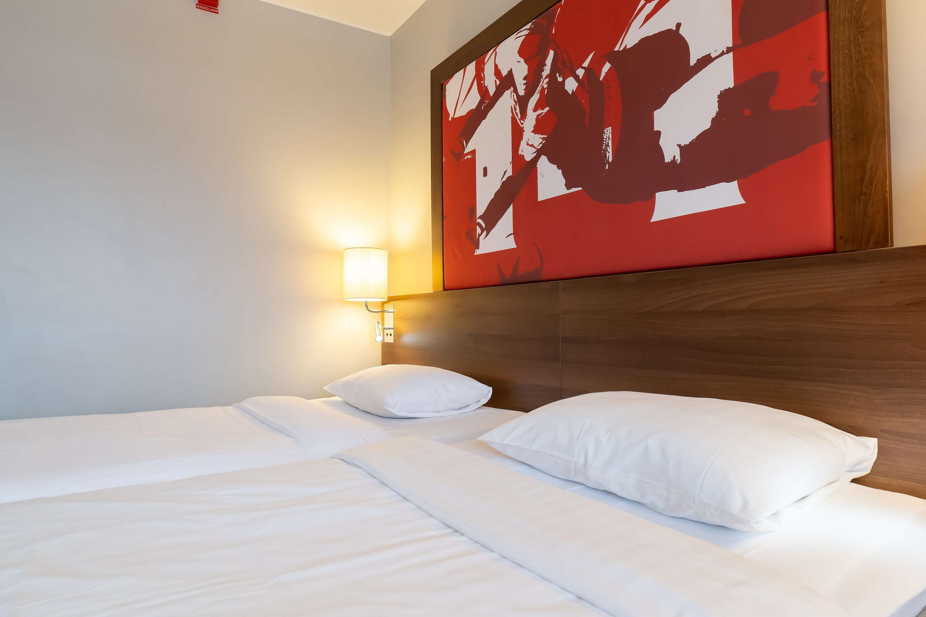 A hotel room with a neatly made bed, a wall-mounted light, and a large abstract painting above the bed.
