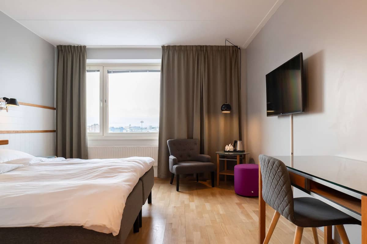 A hotel room with a bed, a chair, a small table with a lamp, a TV, and a window with curtains.