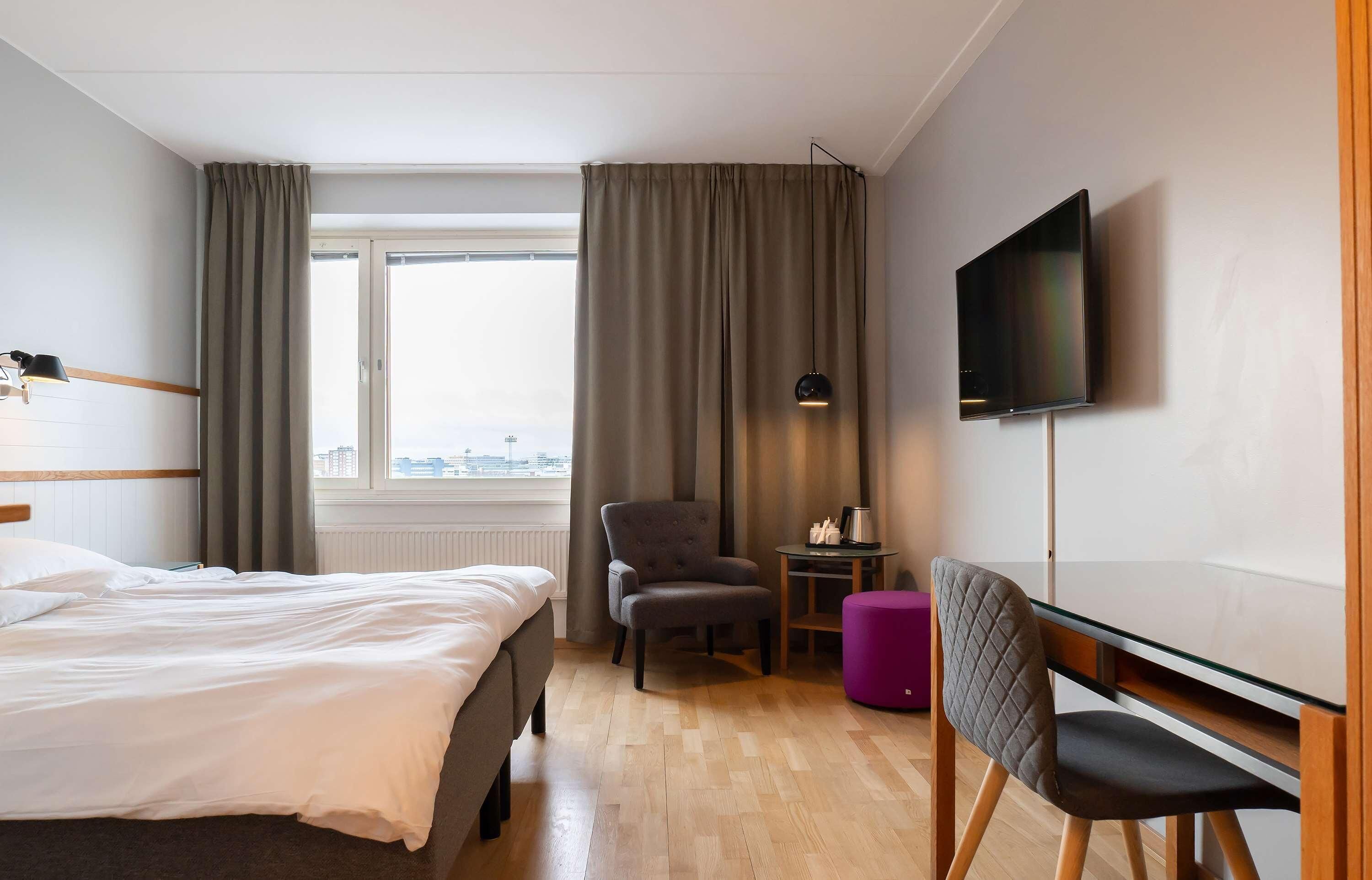Een hotelkamer met een bed, een stoel, een klein tafeltje met een lamp, een televisie en een raam met gordijnen.
