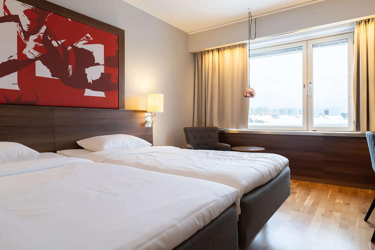 A hotel room with two beds, a large abstract painting, a lamp, a chair, and a view of the city through the window.