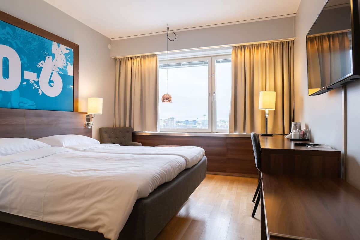 A hotel room with a large bed, a desk, a chair, a TV, and a window with curtains.