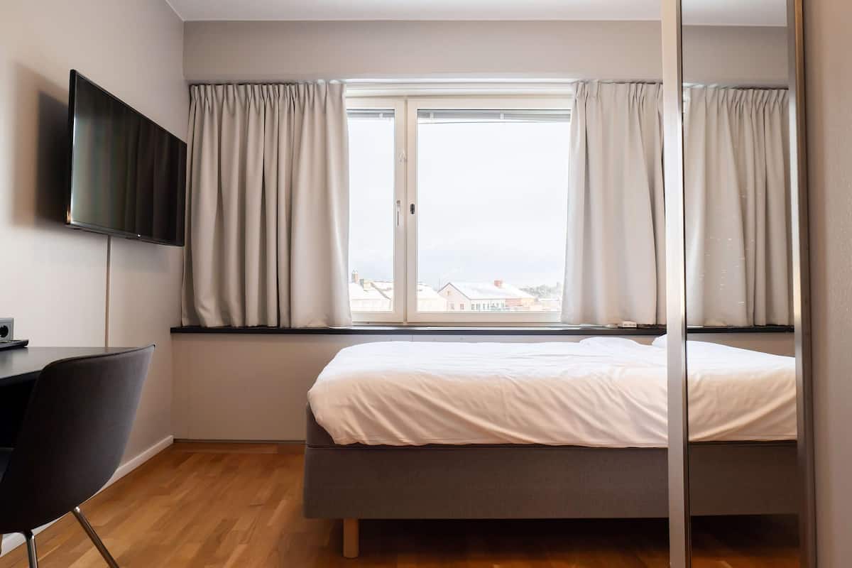 A bedroom with a bed, a TV, a desk, and a window with curtains.