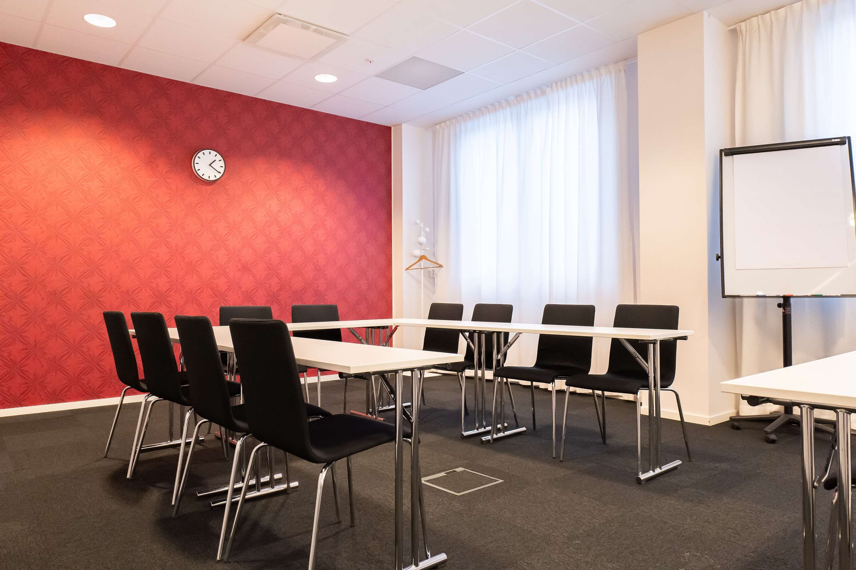 Een congrescentrum met een rode muur, een klok, een whiteboard en zwarte stoelen rond een tafel.