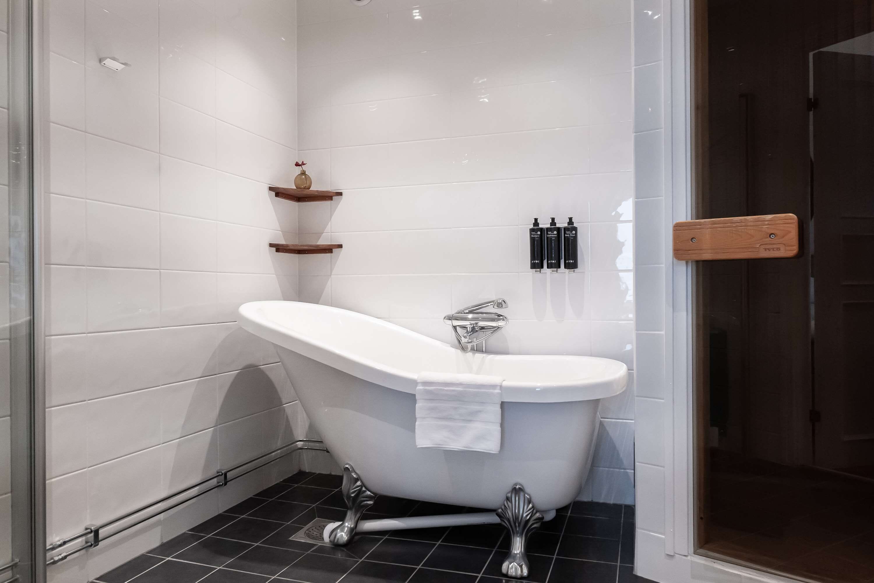 A modern bathroom with a freestanding bathtub, white tiles, a wooden shelf, and a door with a wooden handle.