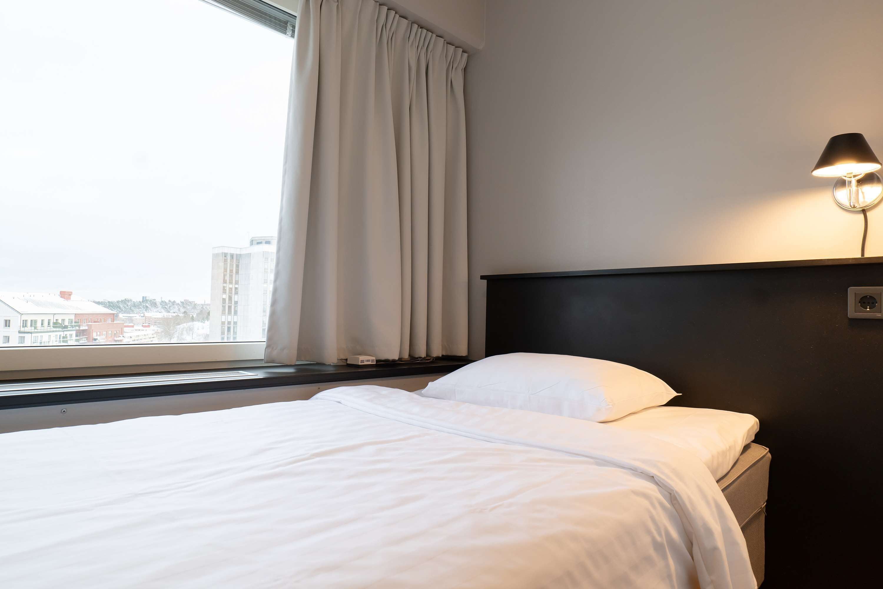 Una camera d'hotel con un letto, un comodino, una lampada, una finestra con tende e vista sugli edifici.