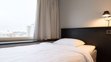 Una camera d'hotel con un letto, un comodino, una lampada, una finestra con tende e vista sugli edifici.