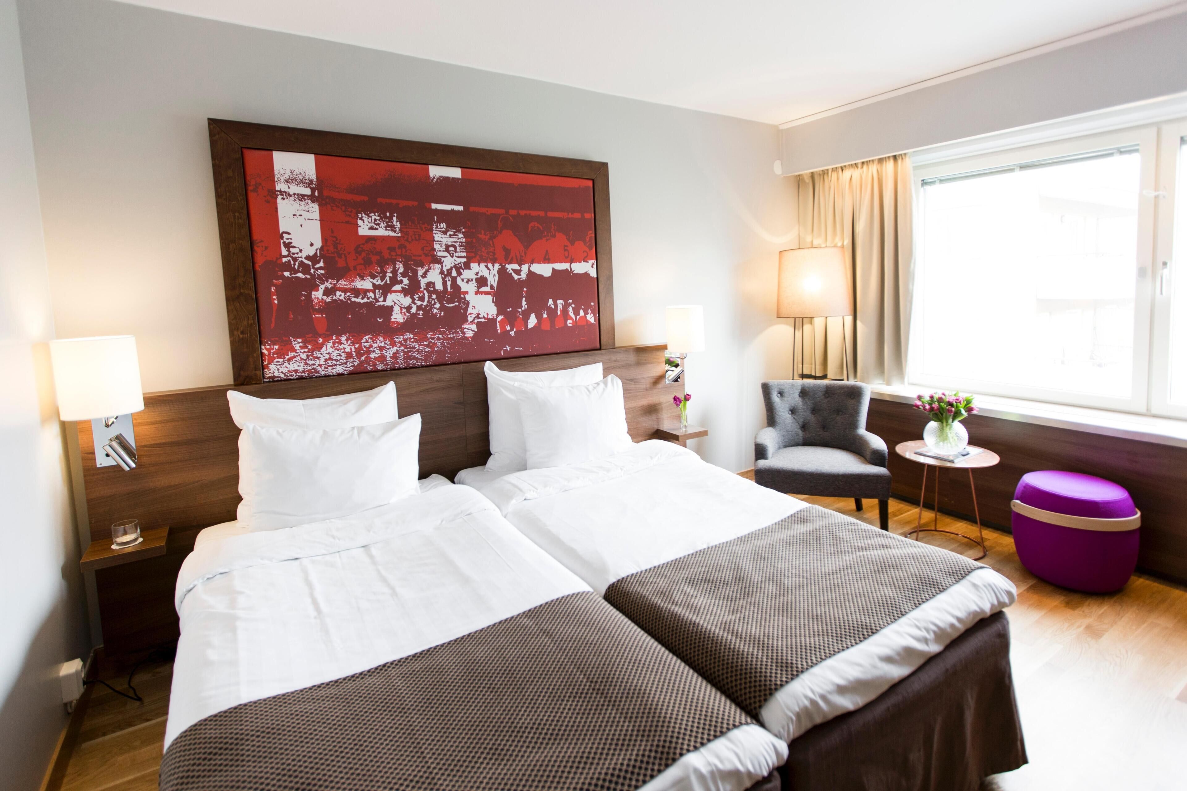 Een hotelkamer met een groot bed, twee nachtkastjes, een stoel, een klein rond tafeltje en een raam met gordijnen.