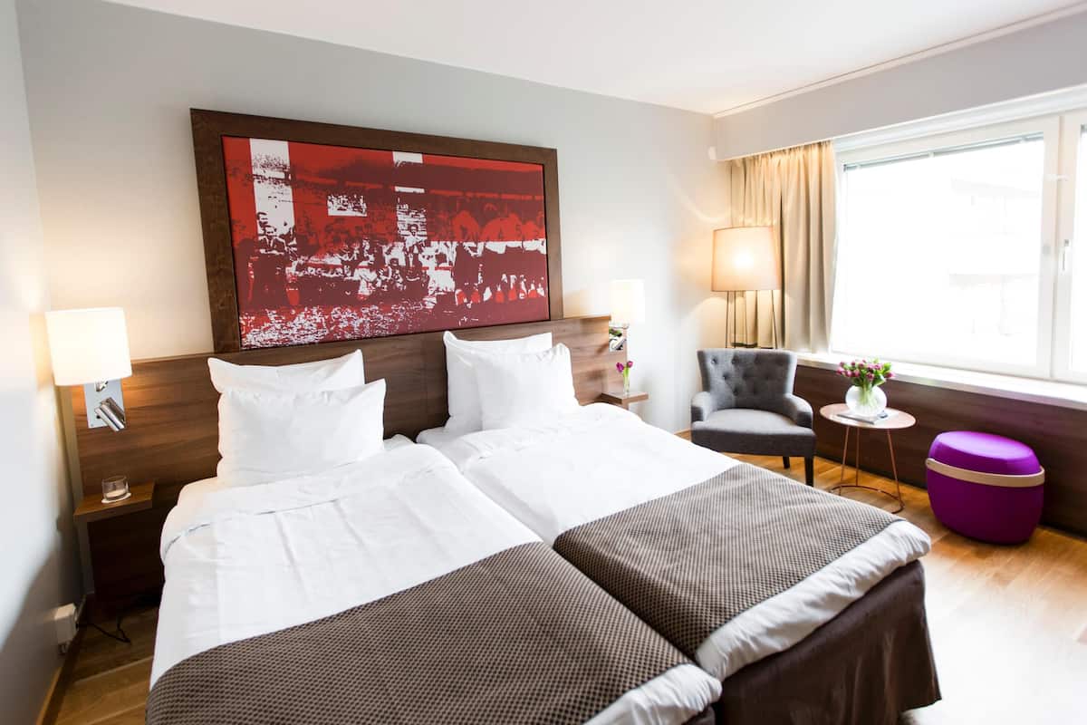 Une chambre d’hôtel avec un grand lit, deux tables de chevet, une chaise, une petite table ronde et une fenêtre avec des rideaux.
