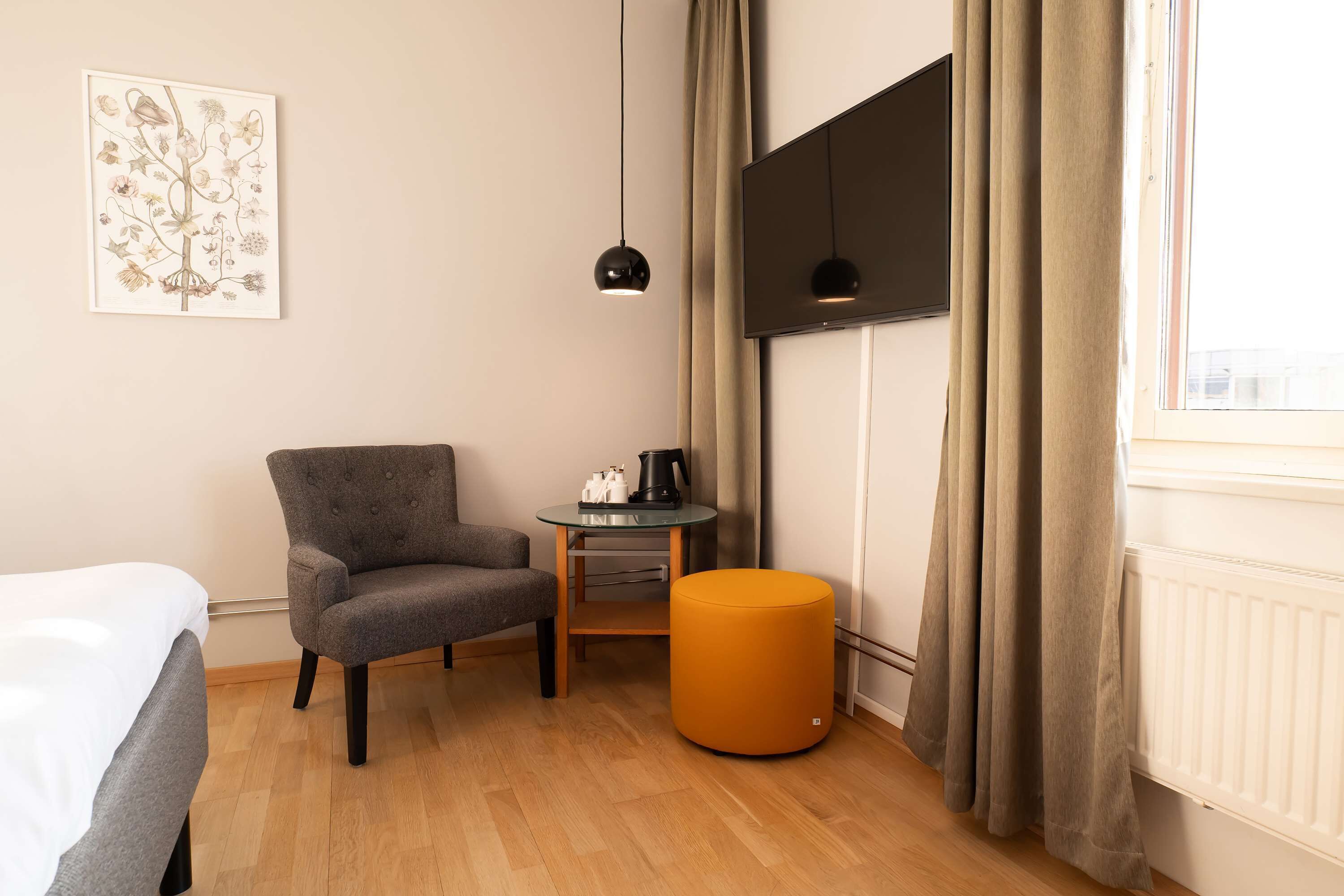 Una habitación de hotel con una cama, un sillón gris, una pequeña mesa redonda con cafetera, un reposapiés naranja y un amplio ventanal con cortinas.