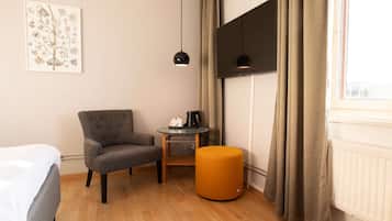 Una habitación de hotel con una cama, un sillón gris, una pequeña mesa redonda con cafetera, un reposapiés naranja y un amplio ventanal con cortinas.