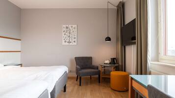 Une chambre d’hôtel avec un lit, un fauteuil gris, une petite table avec une bouilloire et un téléviseur fixé au mur.