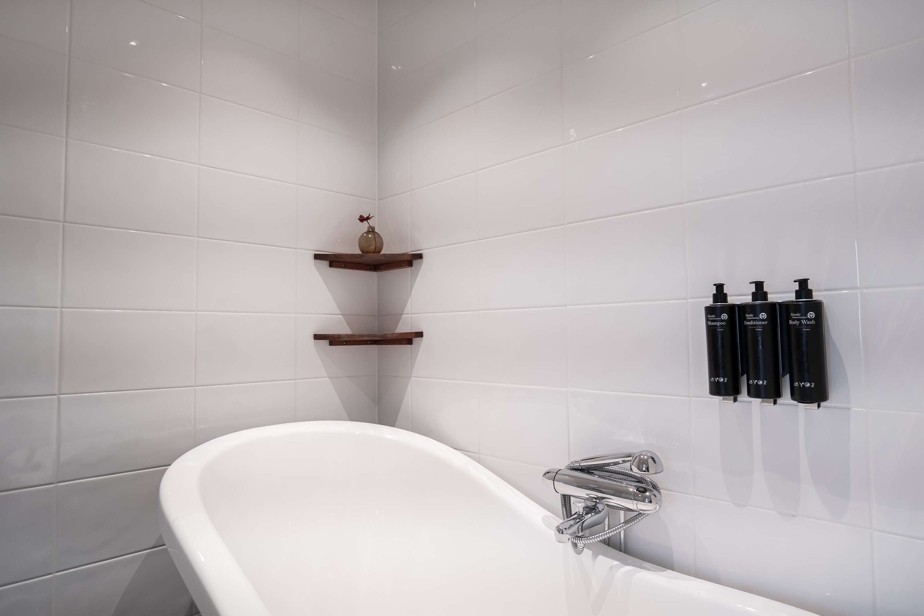 Un baño moderno con una bañera independiente, paredes de azulejos blancos y un estante de madera con un florero.