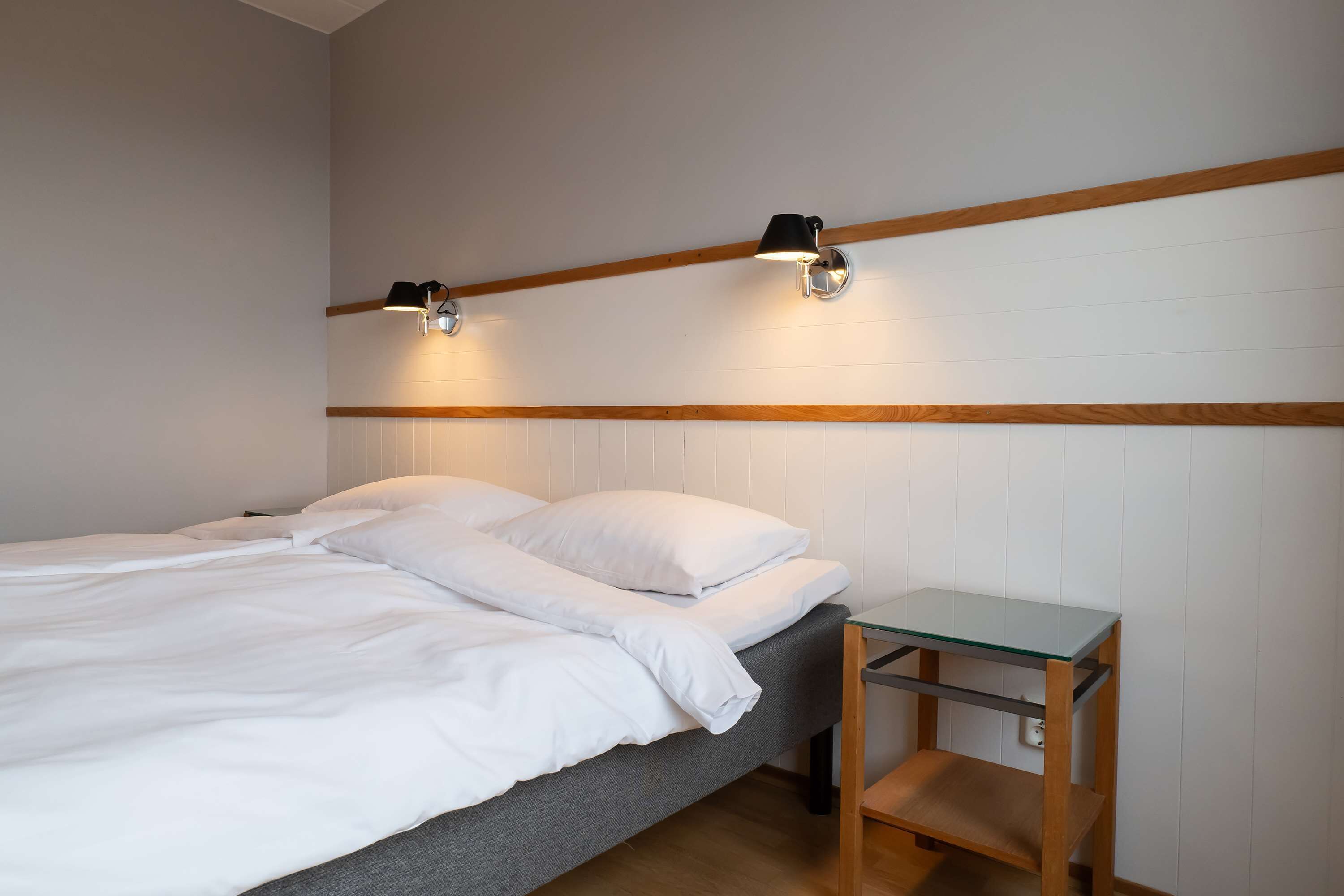 A neatly made bed with white linens, a small wooden nightstand with a glass top, and two wall-mounted lamps.