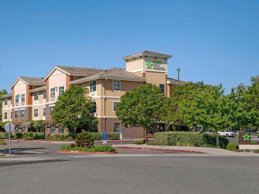 A multi-story hotel with a Stay America sign, surrounded by greenery and a clear sky.