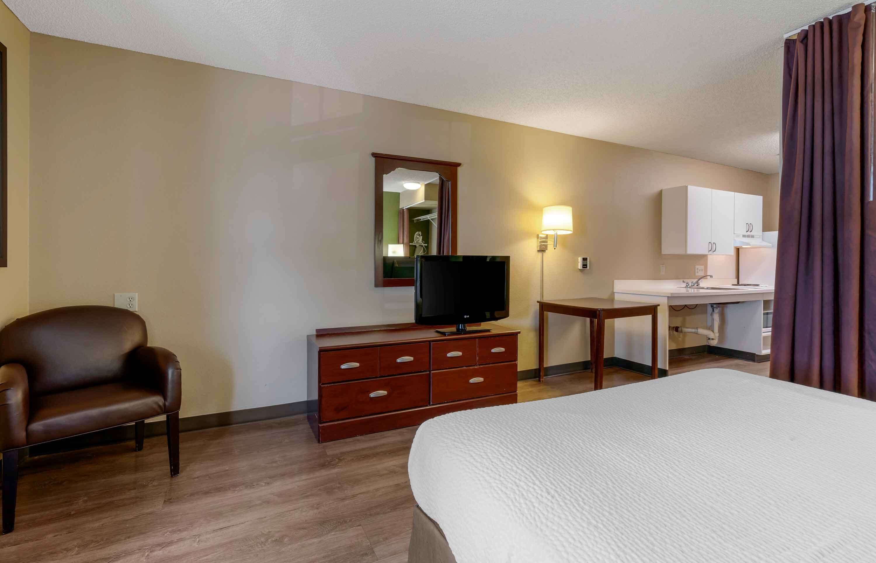 A hotel room with a bed, a TV on a wooden dresser, a desk with a lamp, and a mirror on the wall.