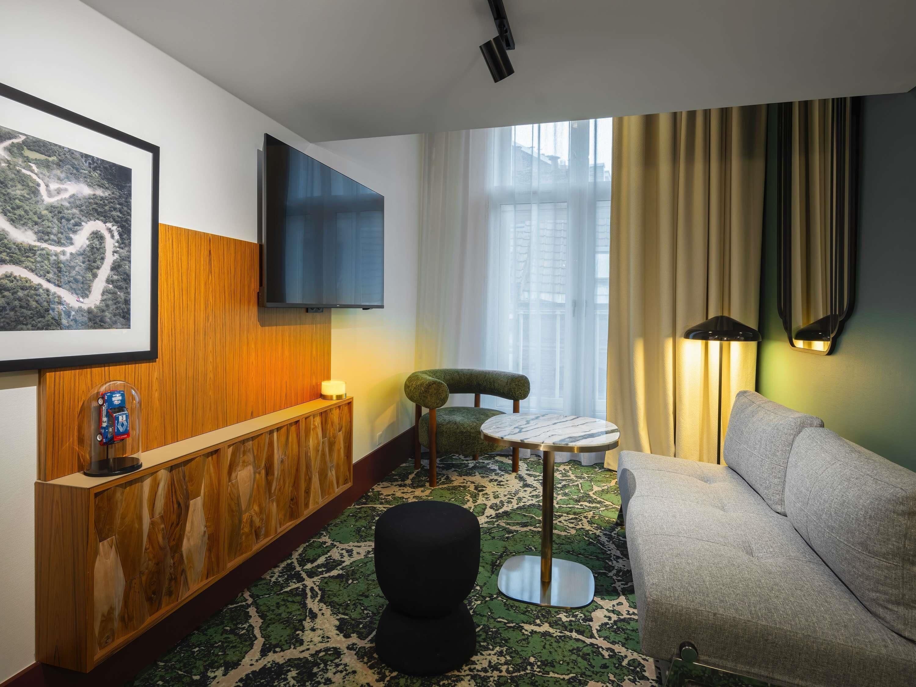 Ein modernes Hotelzimmer mit einem Holzerrezeptionstresen, einem Flachbildfernseher, einem teppichboden in Grünem Muster, einem runden Tisch, einem grünen Sessel, einem grauen Sofa und einem großen Fenster mit Vorhängen.