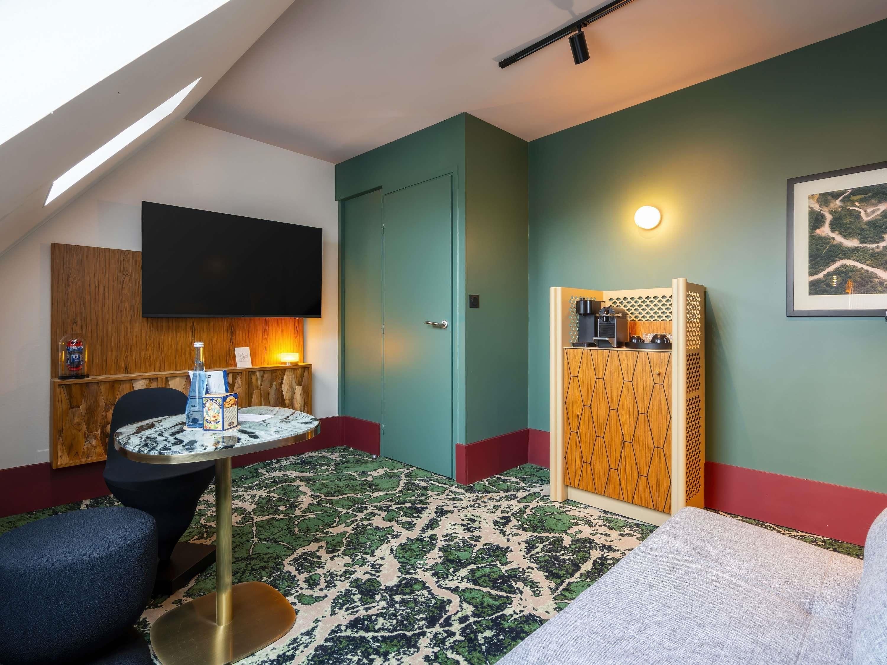 A room with a TV mounted on the wall, a small table with a bottle and a menu, a sofa, and a door leading to another room.