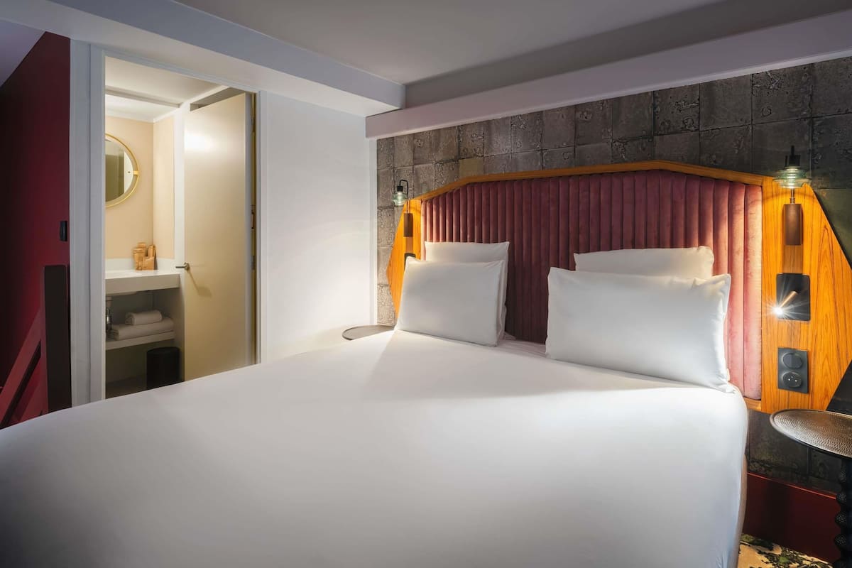 Une chambre d’hôtel avec un grand lit, une tête de lit en bois, une table de chevet avec une lampe, et une salle de bain visible à travers une porte ouverte.