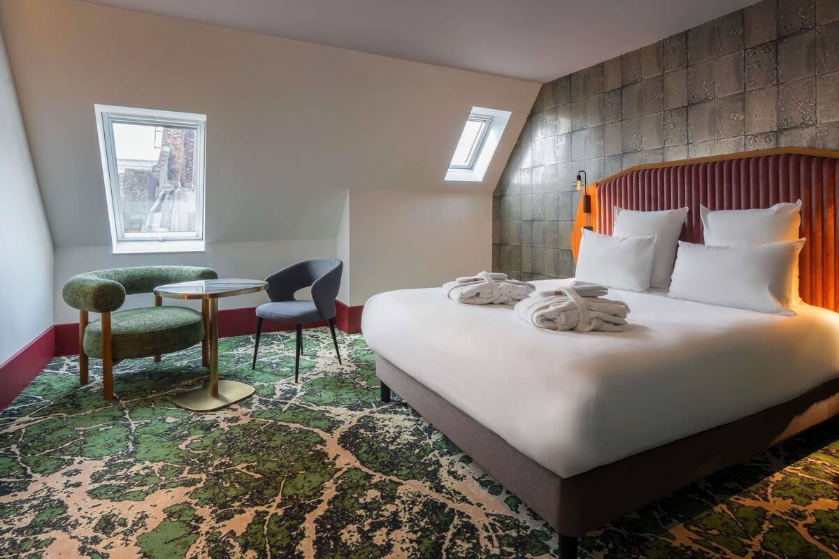 Une chambre d’hôtel avec un grand lit, deux fauteuils, une petite table et une vue sur la ville grâce à une lucarne.