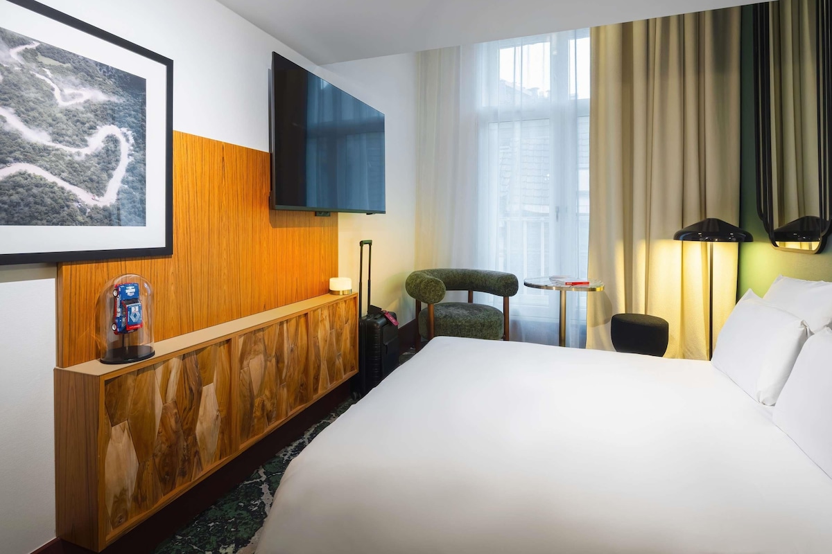 Una habitación de hotel con una cama grande, una consola de madera, un televisor montado en la pared, una silla, una mesita y una lámpara.