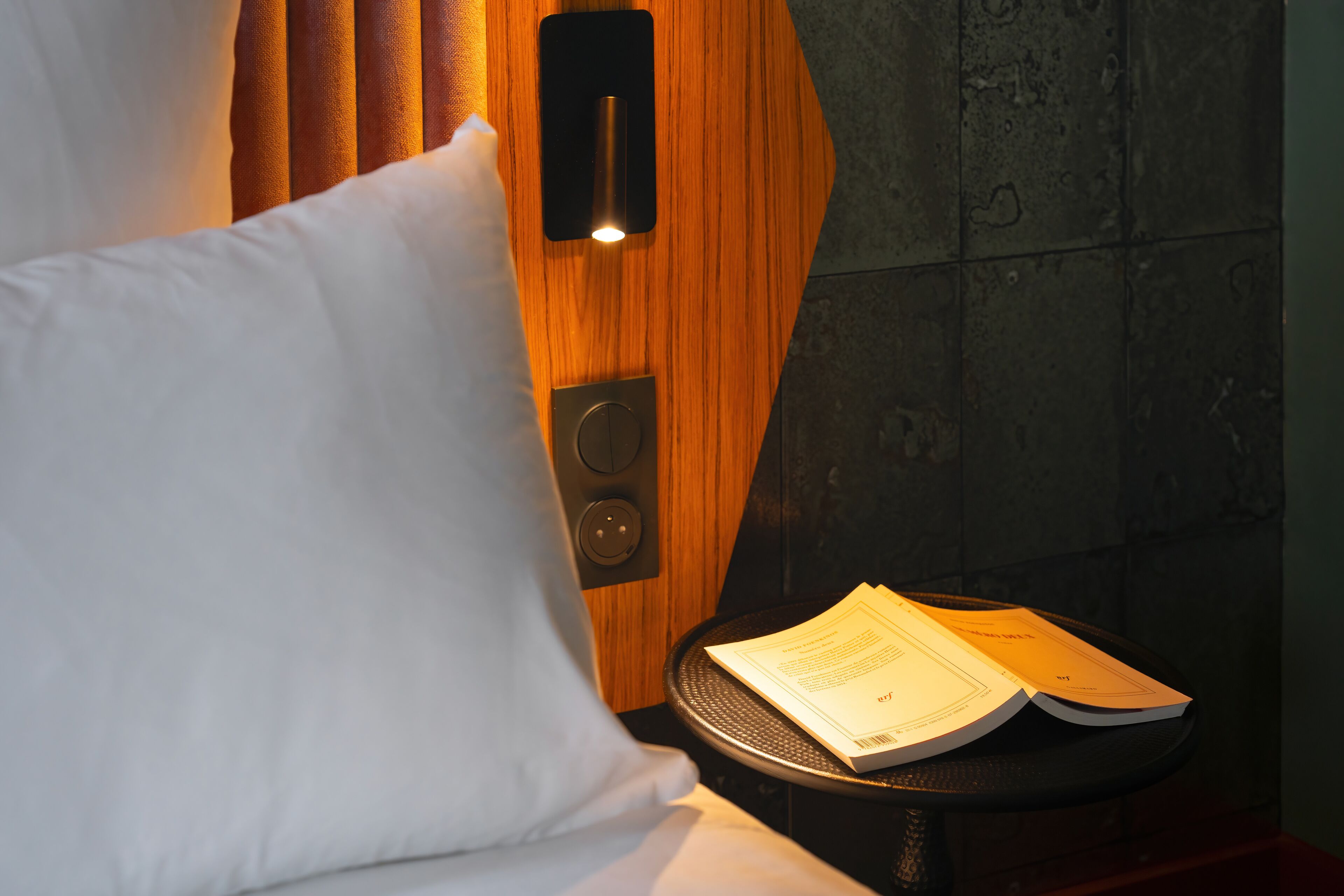 Ein Hotelzimmer mit einem hölzernen Kopfteil, einem weißen Kissen und einem kleinen Tisch mit einem Buch.