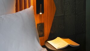 Een hotelkamer met een houten hoofdeinde, een wit kussen en een tafeltje met een boek.
