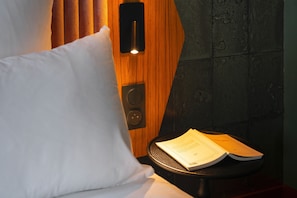 Una habitación de hotel con cabecera de madera, almohada blanca y una pequeña mesa con un libro.