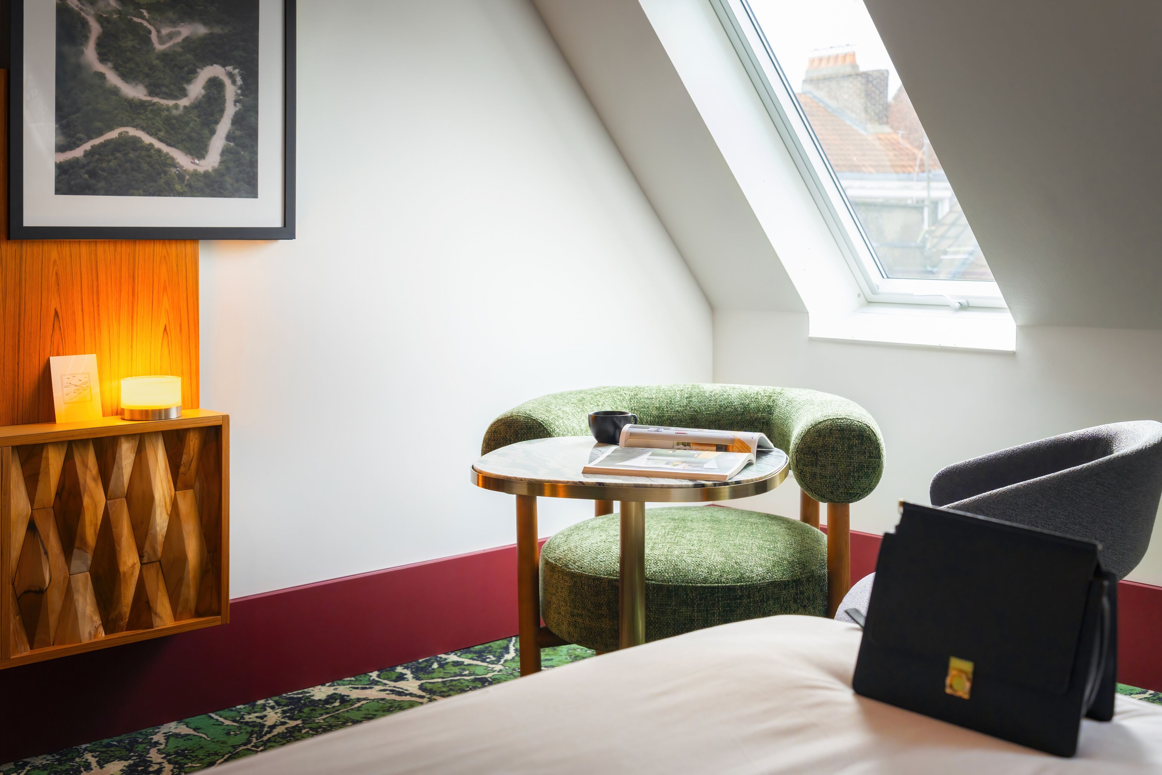 A room with a bed, a green upholstered chair, a small round table, a framed picture on the wall, and a window with a view of buildings.
