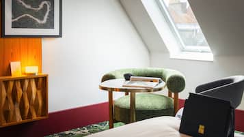 A room with a bed, a green upholstered chair, a small round table, a framed picture on the wall, and a window with a view of buildings.