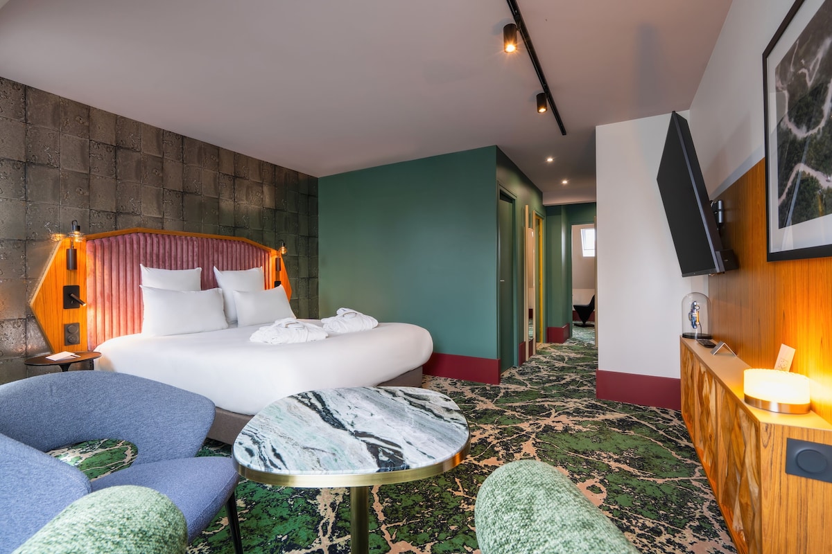 Una habitación de hotel con una cama grande, una mesa redonda de mármol, un sillón azul y una recepción.