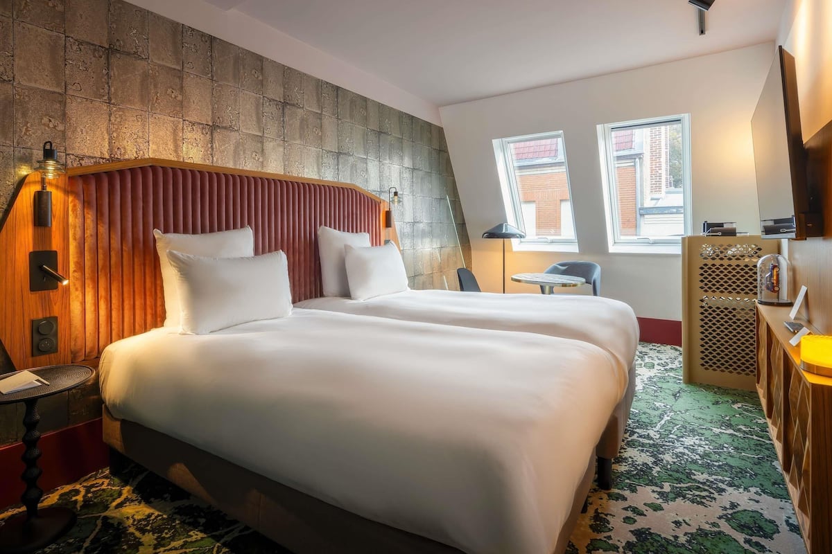 Une chambre d’hôtel avec un grand lit, un bureau, une chaise et une petite table. Il y a deux fenêtres et un luminaire au plafond.