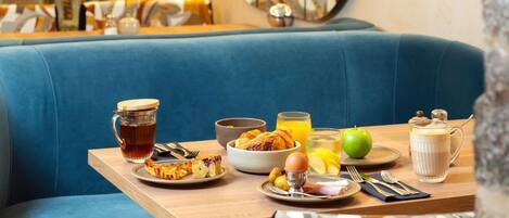A breakfast setup on a wooden table with a blue cushioned bench, a glass pitcher, a bowl of fruit, a plate of eggs, and a glass of orange juice.