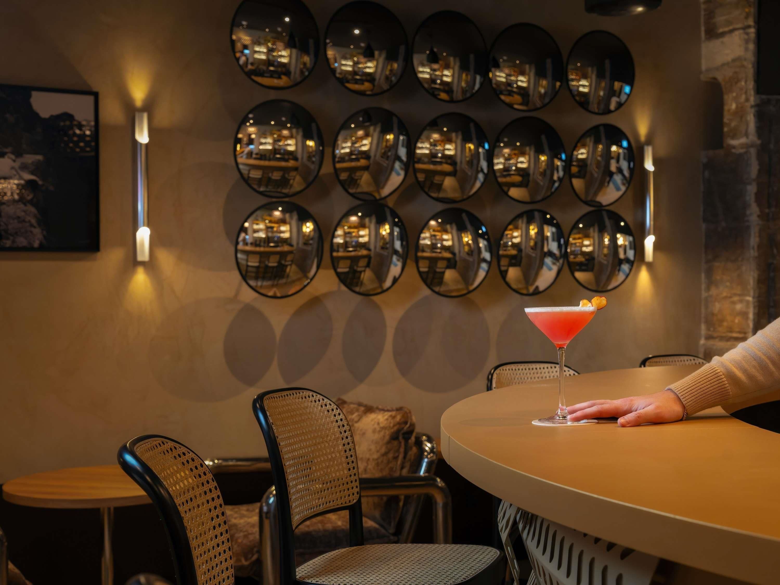A bar area with multiple round mirrors on the wall, a table with a single cocktail glass, and two wicker chairs.
