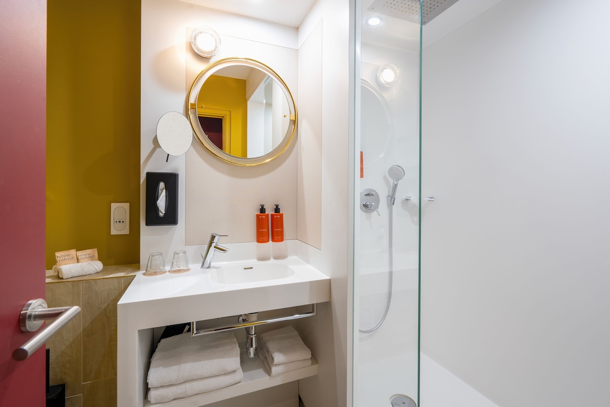 Un baño moderno con un lavamanos blanco, un espejo redondo y una ducha con mampara de vidrio.