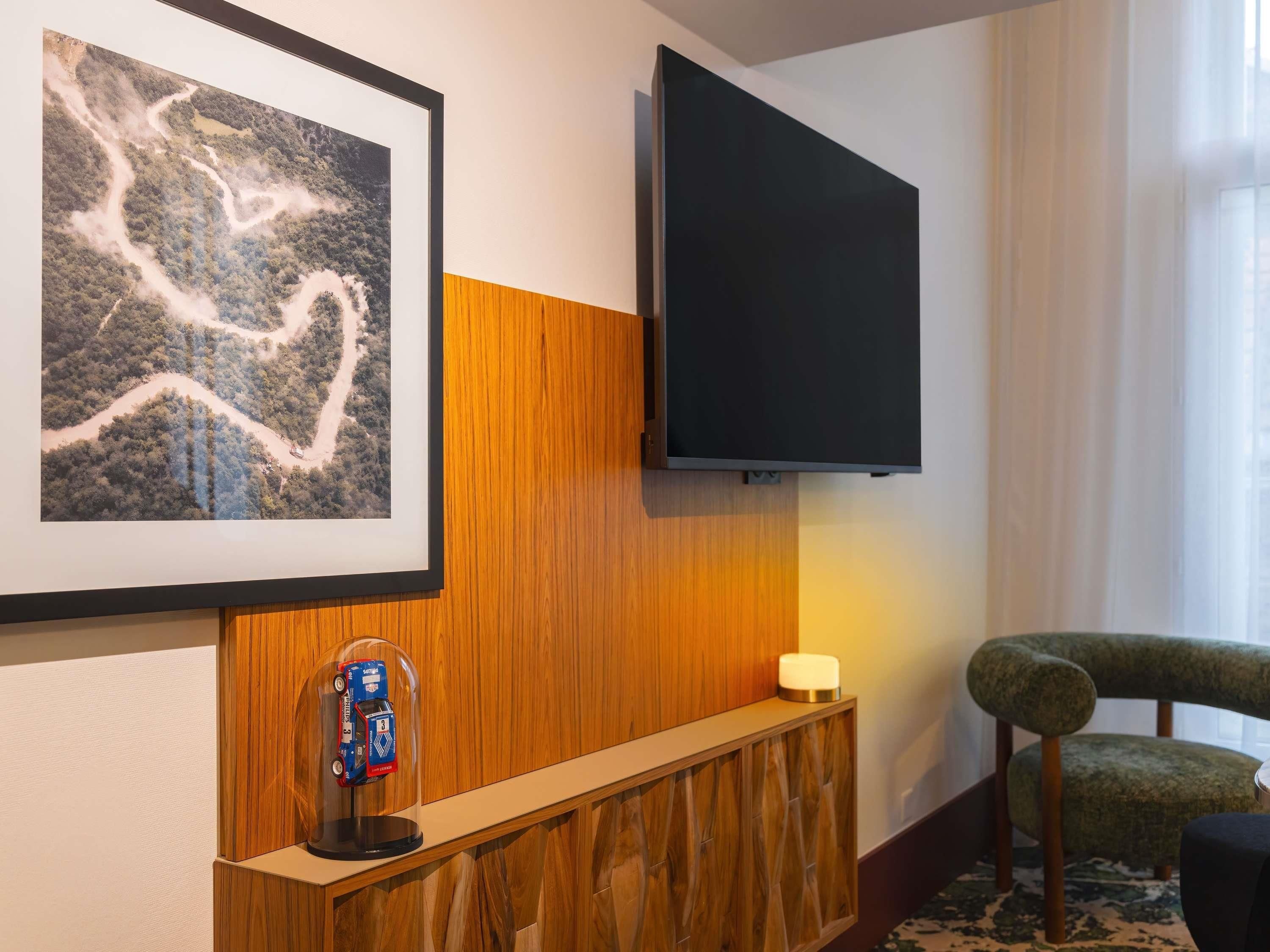 A hotel room with a large framed landscape painting, a mounted flat-screen TV, a wooden console table, a small toy train, and a green upholstered chair.