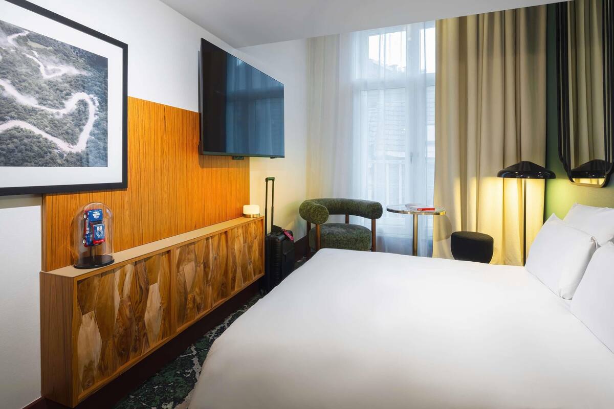 Een hotelkamer met een groot bed, een houten console, een aan de muur bevestigde televisie, een stoel, een klein tafeltje en een lamp.