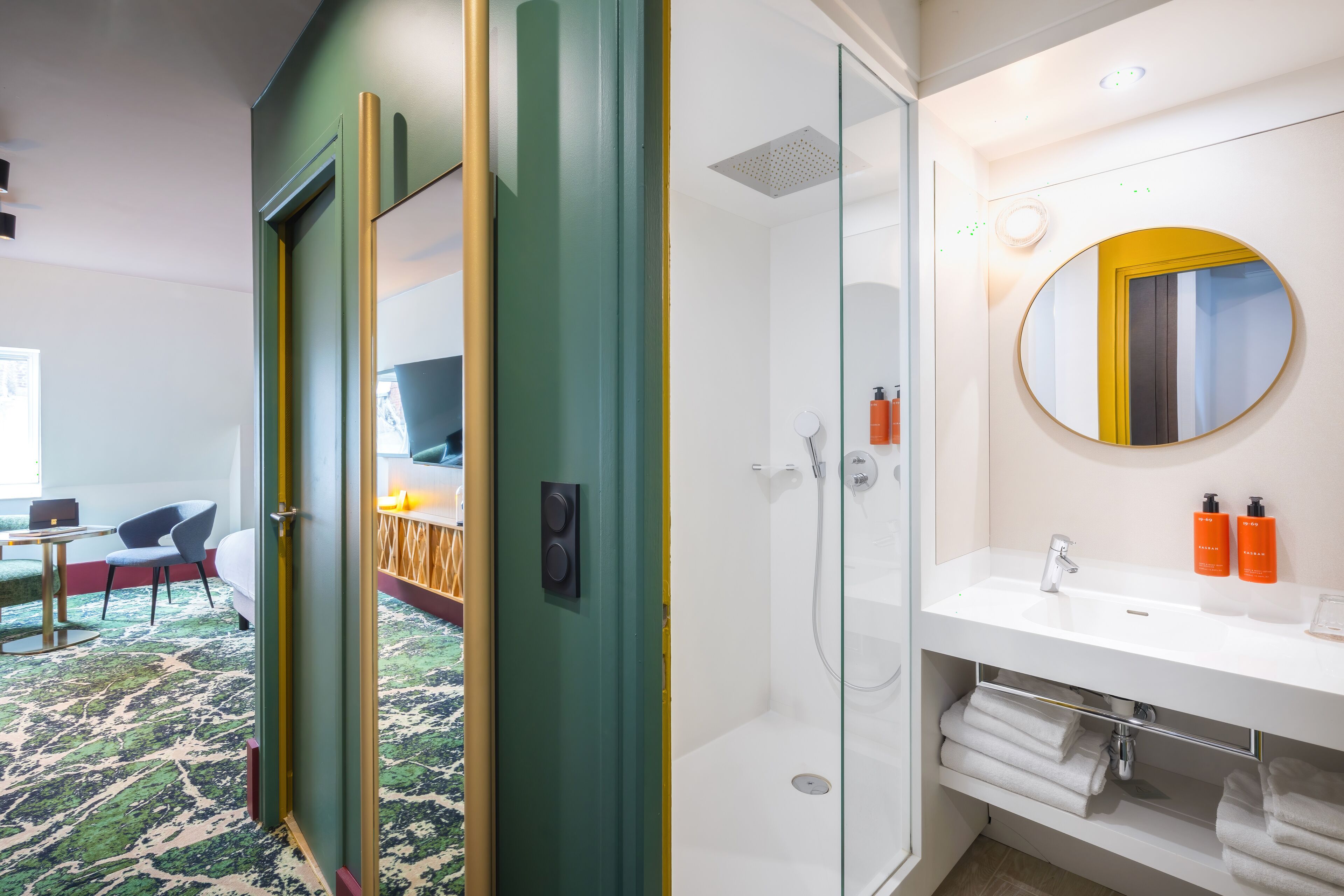 A hotel room with a glass-enclosed shower, a white sink with a mirror, and a bed with a green door.