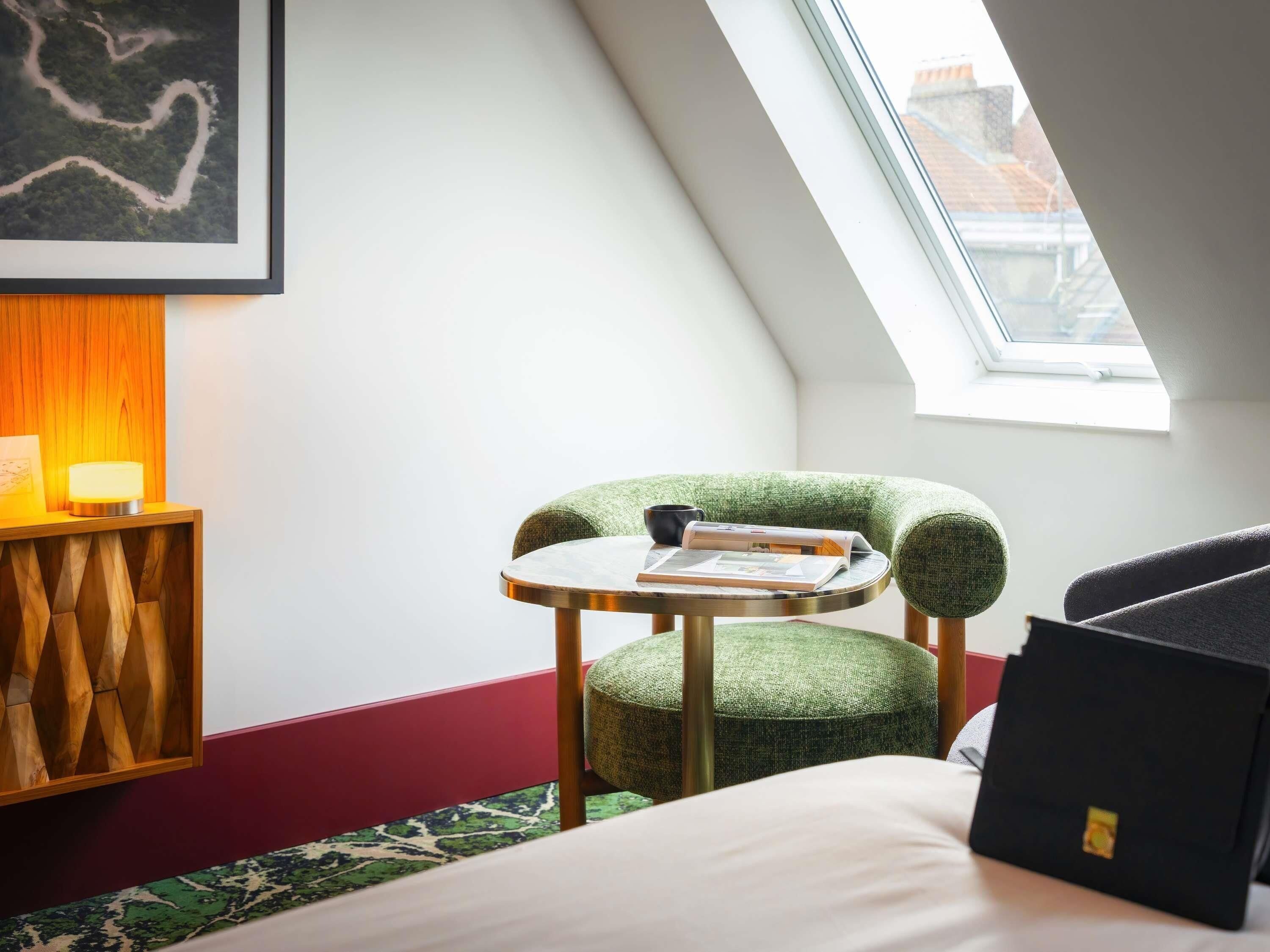 Een kamer met een schuine kap, een groene beklede stoel, een klein rond tafeltje, een bed en een lijst met een afbeelding aan de muur.
