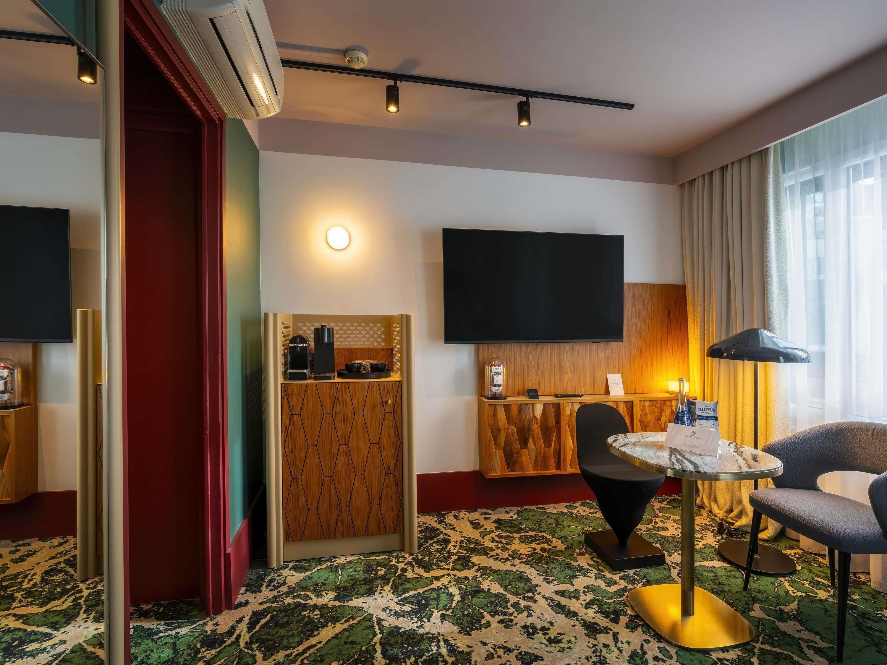 Une chambre d’hôtel dotée d’un grand téléviseur à écran plat, d’une petite kitchenette, d’une table ronde avec des chaises et d’un tapis à motifs.